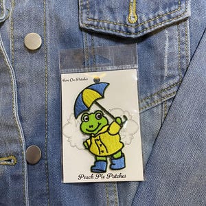 Puede incluir: Parche para planchar con una rana verde que lleva un impermeable amarillo y sostiene un paraguas azul y amarillo. El parche está en una tarjeta blanca con el texto "Iron On Patches" y "Peach Pie Patches".