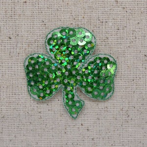 May include: A green sequined shamrock applique with a silver border. The three-leaf clover is filled with shiny green sequins. The applique is on a beige fabric background. Perfect for St. Patrick's Day.