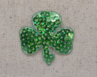 May include: A green sequined shamrock applique with a silver border. The three-leaf clover is filled with shiny green sequins. The applique is on a beige fabric background. Perfect for St. Patrick's Day.