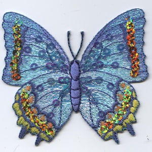 以下が含まれることがあります： 金色とオレンジ色のスパンコールが付いた、虹色の青い蝶のアップリケ。蝶は細部まで刺繍されており、キラキラとした仕上がりになっています。