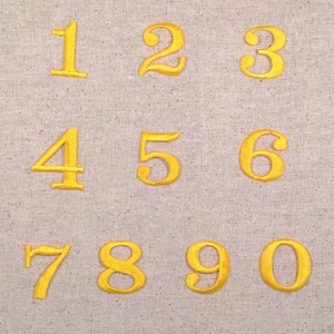 May include: A set of ten gold embroidered numbers, 1 through 10, on a beige fabric background.