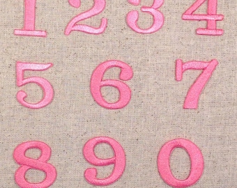 May include: Pink felt numbers 1 to 0 on a beige fabric background.