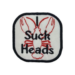 Op de afbeelding: Geborduurde patch met een witte achtergrond, zwarte rand en de woorden "I Suck Heads" in zwart en rood. Het ontwerp toont een rode kreeftenillustratie.