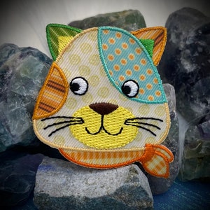 May include: A colorful patchwork cat face applique with a yellow nose, brown eyes, and a yellow and orange striped scarf. The applique is made of various fabrics with different patterns, including polka dots, stripes, and a floral design.
