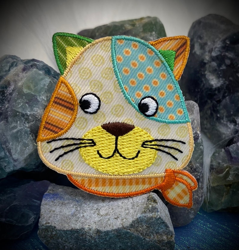May include: A colorful patchwork cat face applique with a yellow nose, brown eyes, and a yellow and orange striped scarf. The applique is made of various fabrics with different patterns, including polka dots, stripes, and a floral design.