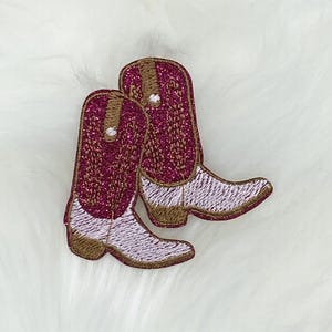 May include: A pair of pink and white cowboy boot patches with gold glitter accents. The boots are embroidered with a detailed design.