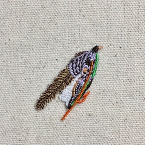 Puede incluir: Parche bordado de un pájaro morado y marrón con detalles verdes y naranjas. El pájaro está posado en una rama con las alas extendidas.