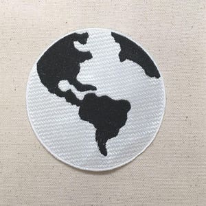 May include: A round, white embroidered patch featuring a black and white world map design. The continents are depicted in black against a white background. The patch is circular and has a detailed, textured appearance.