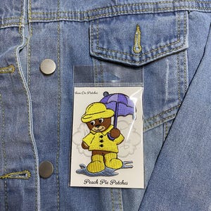 Puede incluir: Parche para planchar con un oso amarillo que lleva un impermeable y sostiene un paraguas morado. El parche está en una tarjeta blanca con el texto "Iron On Patches" y "Peach Pie Patches".