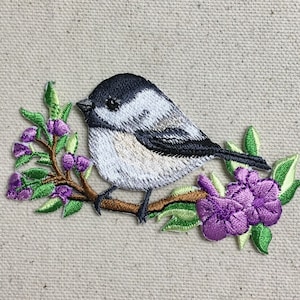 Puede incluir: Parche bordado para planchar con un pequeño pájaro blanco y negro posado en una rama con flores moradas. El pájaro está mirando hacia la izquierda y tiene un pequeño pico negro.