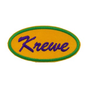 Puede incluir: Parche bordado con la palabra "Krewe" en escritura morada sobre un óvalo amarillo. El óvalo tiene un borde verde. Este parche está diseñado para aplicarse en ropa o accesorios.