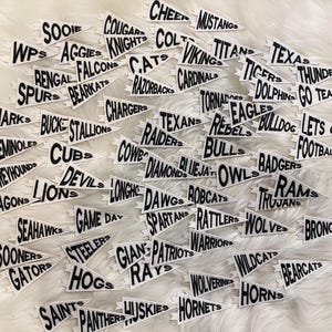 Puede incluir: Una colección de banderines blancos con texto negro, cada uno con el nombre de un equipo deportivo diferente. Los banderines están dispuestos en un patrón aleatorio sobre una superficie blanca.