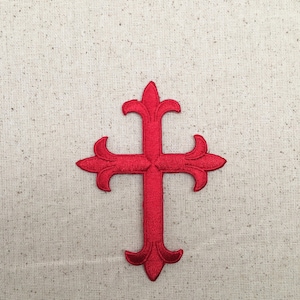 Puede incluir: Una cruz roja bordada con un diseño decorativo. La cruz está sobre un fondo de tela beige.