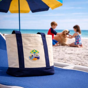 May include: A canvas tote bag with navy blue accents and handles, featuring an embroidered beach scene with an umbrella, food, and a cooler. The bag is on a blue lounge chair, with a beach and ocean in the background.