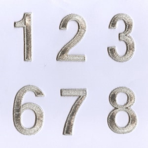 May include: Ten silver embroidered iron-on numbers, 0 through 9, on a white background.