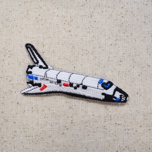 May include: White and black embroidered patch of a space shuttle with blue and red details.