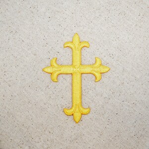 Könnte beinhalten: Ein golden besticktes Kreuz mit einem einfachen Design, bestehend aus einem Mittelpunkt und vier Armen mit abgerundeten Enden. Das Kreuz befindet sich auf einem beigefarbenen Stoffhintergrund.