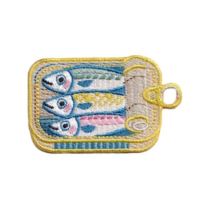 Puede incluir: Parche bordado con tres peces coloridos dentro de una lata de sardinas abierta. Los peces tienen detalles azules, amarillos y rosas, con ojos azules y blancos. La lata es beige con un borde y una anilla amarillos.