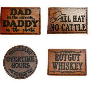 May include: Four brown leather patches with black text. The first patch says "DAD in the streets, DADDY in the sheets". The second patch says "ALL HAT NO CATTLE". The third patch says "WORKING ALL DAY OVERTIME HOURS FOR BULLSHIT PAY". The fourth patch says "ROTGUT WHISKEY".
