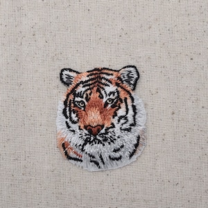 Puede incluir: Parche bordado para planchar con la cara de un tigre. El tigre tiene rayas naranjas y negras y un hocico blanco.