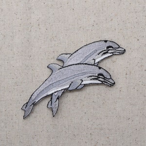 May include: Two gray embroidered dolphin patches, one on top of the other, with the bottom dolphin's tail extending to the left and the top dolphin's tail extending to the right.