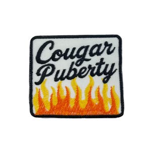 Może przedstawiać: Haftowana naszywka z napisem "Cougar Puberty" czarną czcionką na białym tle. Naszywka przedstawia płomienie w odcieniach żółci i pomarańczy na dole, z czarną obwódką.