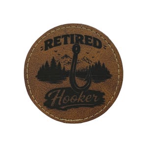 以下が含まれることがあります： 黒い文字で「RETIRED Hooker」と書かれた丸い茶色の革パッチ。デザインには、黒い釣り針、木、山が含まれています。パッチにはステッチされたボーダーがあります。