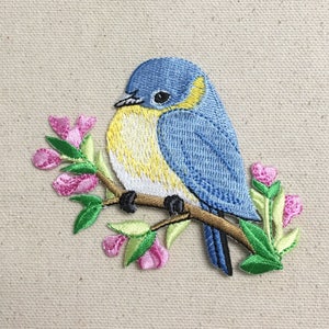 May include: Embroidered iron-on patch featuring a bluebird perched on a branch with pink flowers. The bird has a yellow breast and a blue head.