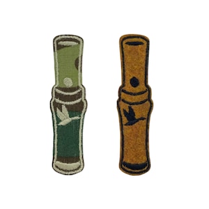 May include: Two embroidered patches in the shape of duck calls. One is green and brown with a white duck graphic. The other is tan with a black duck graphic. Both have a rounded top and bottom.