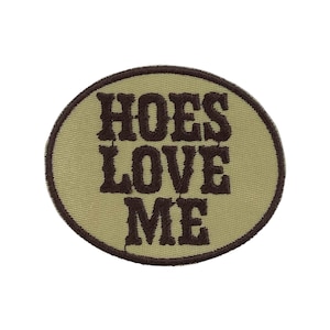 以下が含まれることがあります： ベージュ色の背景に、濃い茶色で「HOES LOVE ME」と刺繍された楕円形のパッチ。 パッチは茶色の縁取りがあり、衣服やアクセサリーに縫い付けるように設計されています。