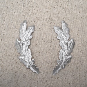 May include: Two silver-coloured fabric leaf embellishments, each with a curved shape and a design of overlapping leaves. The embellishments are on a light beige fabric background.