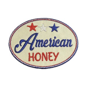 May include: Oval embroidered patch with the words "American HONEY" in blue and red script. The patch features three stars, a red, a white, and a blue star, above the word "American". The patch has a cream-colored background and a red and white border.