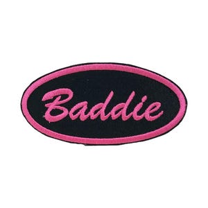 May include: An oval-shaped patch with a black background and a pink border. The word "Baddie" is embroidered in pink cursive script across the center. The patch is likely designed to be sewn or ironed onto clothing or accessories.