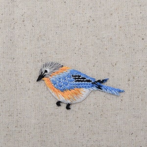 May include: A bluebird embroidered on a beige fabric background. The bird has a blue and white body with orange accents and black eyes.