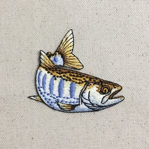 May include: An embroidered patch of a brown and white fish with blue stripes. The fish is facing right and has a yellow tail fin.