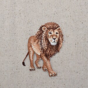 May include: Embroidered lion patch with brown and tan fur, a detailed face, and a long tail. The lion is standing on a beige fabric background.