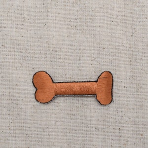 May include: A brown embroidered bone-shaped patch. The patch is on a beige fabric background.