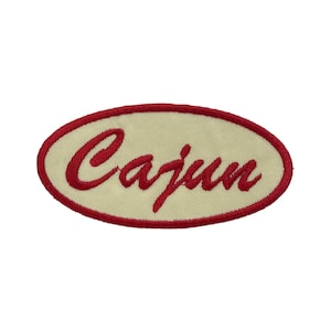 May include: An embroidered patch with the word "Cajun" in red script lettering. The text is centered on a cream-colored oval, framed by a red border. The patch is likely for clothing or accessories.