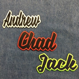 Pode incluir: Três patches para passar a ferro com nomes escritos em uma fonte estilizada. Os nomes são "Andrew" em preto sobre branco, "Chad" em preto sobre vermelho e "Jack" em preto sobre verde.
