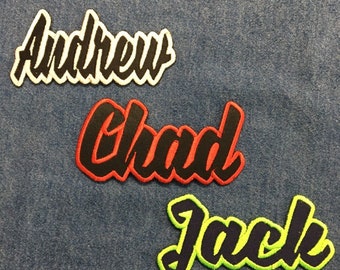 以下が含まれることがあります： 3つの刺繍名札。「Andrew」は白の縁取りのある黒い筆記体、「Chad」は黒の縁取りのある赤い筆記体、「Jack」はネオングリーンの縁取りのあるネイビーの筆記体。パッチは青いデニムの背景にあります。