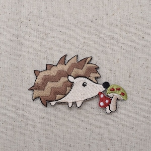 以下が含まれることがあります： 茶色と白の針を持つハリネズミの刺繍パッチ。白い顔と小さな黒い目があります。ハリネズミは2つのキノコを持っており、1つは白い水玉模様の赤色、もう1つは赤い水玉模様の緑色です。パッチはベージュ色の生地の背景にあります。