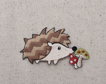 以下が含まれることがあります： 茶色と白の針を持つハリネズミの刺繍パッチ。白い顔と小さな黒い目があります。ハリネズミは2つのキノコを持っており、1つは白い水玉模様の赤色、もう1つは赤い水玉模様の緑色です。パッチはベージュ色の生地の背景にあります。