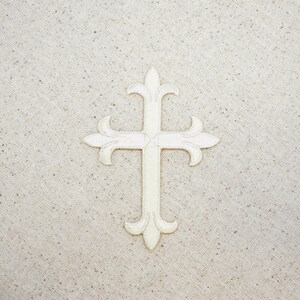 May include: A white embroidered cross with a decorative design on a beige fabric background.