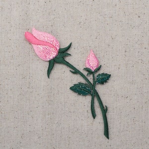 May include: A pink embroidered rose with a bud, with green leaves and stems, on a beige fabric background.