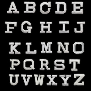 May include: A complete set of twenty-six white embroidered alphabet letters from A to Z arranged in five rows against a solid black background. Each letter features a bold, block-style font with a textured, stitched appearance and clean edges.