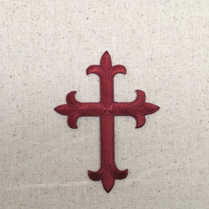 May include: A burgundy embroidered cross with a decorative design. The cross is on a white linen background.