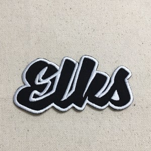 May include: A black and white embroidered patch featuring the word "elles" in a cursive font. The patch has a black base with white stitching outlining the letters and border. The patch is set against a beige fabric backdrop.