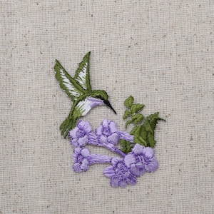 Può includere: Colibrì ricamato verde e bianco con un lungo becco, posato su un gruppo di fiori viola con foglie verdi.
