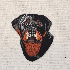 May include: Embroidered patch of a Rottweiler dog's head. The dog has black fur with brown markings around the eyes and mouth.