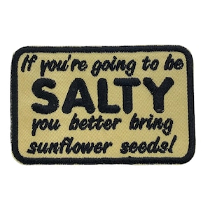 以下が含まれることがあります： タン色の背景に黒い文字で「If you're going to be SALTY you better bring sunflower seeds!」と書かれた刺繍パッチ。パッチは黒い縁取りがあり、長方形です。